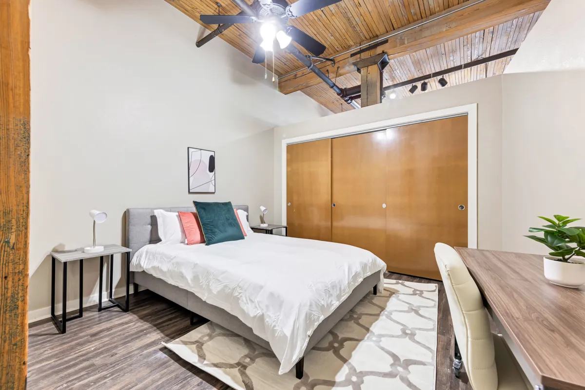 The image shows a sunlit bedroom featuring a plush bed with crisp white bedding and colorful accent pillows, set beneath striking exposed wooden beams. A sleek desk with a modern chair and a touch of greenery sits nearby, creating a perfect nook for work or reflection. The overall atmosphere is calm, inviting, and infused with historic character.