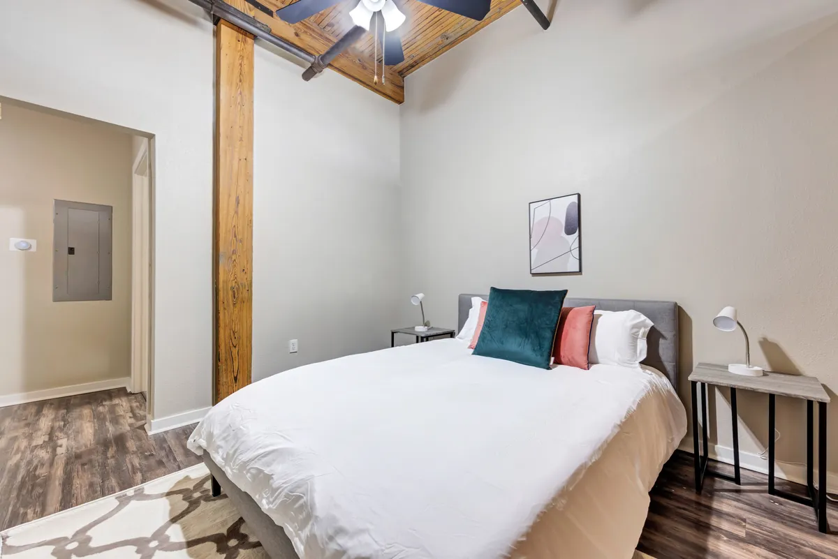 A cozy bedroom scene featuring a comfortable bed adorned with inviting blankets, ceiling fan overhead, tasteful artwork, and nightstands on either side. Sunlight filters through, enhancing the room’s warmth and tranquility.