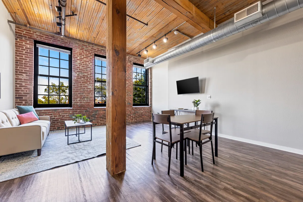 This image showcases a spacious living area with a large sofa, a dining table, huge windows streaming in natural light, exposed brick walls, and rustic wooden beams. The open-plan layout is evident, with plush furnishings and architectural details blending seamlessly. The natural light illuminates the space, emphasizing its inviting and historic character.