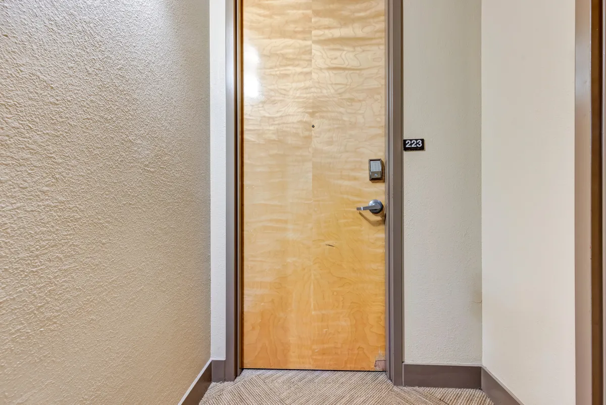 A close-up view of a wooden door with a distinct door number, set within a simple but inviting hallway. The image hints at a welcoming entry point, suggesting the transition from inside the building to the surrounding urban environment.