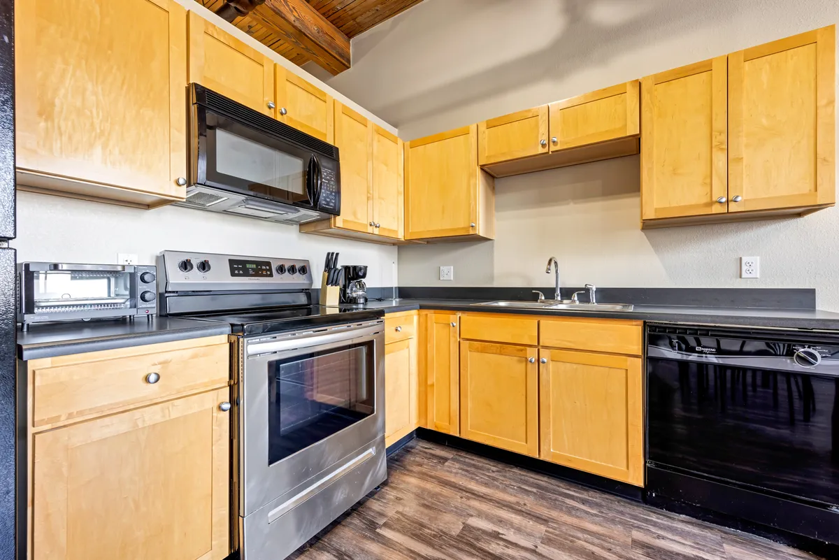 A modern kitchen space featuring stainless steel appliances, warm wooden cabinets, and a sleek black countertop. The inviting setup hints at opportunities for cooking, gathering with friends, and entertaining in a comfortable, home-like environment.