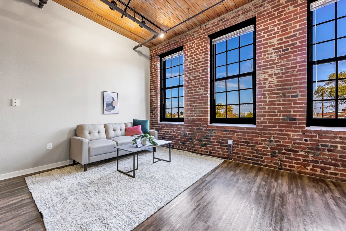 A cozy living lounge featuring a comfortable sofa and coffee table set beside large windows. Exposed brick walls provide a dramatic, historic backdrop, while the soft furnishings and ample sunlight give the space a welcoming, relaxing ambiance. The open area invites both socializing and quiet moments.