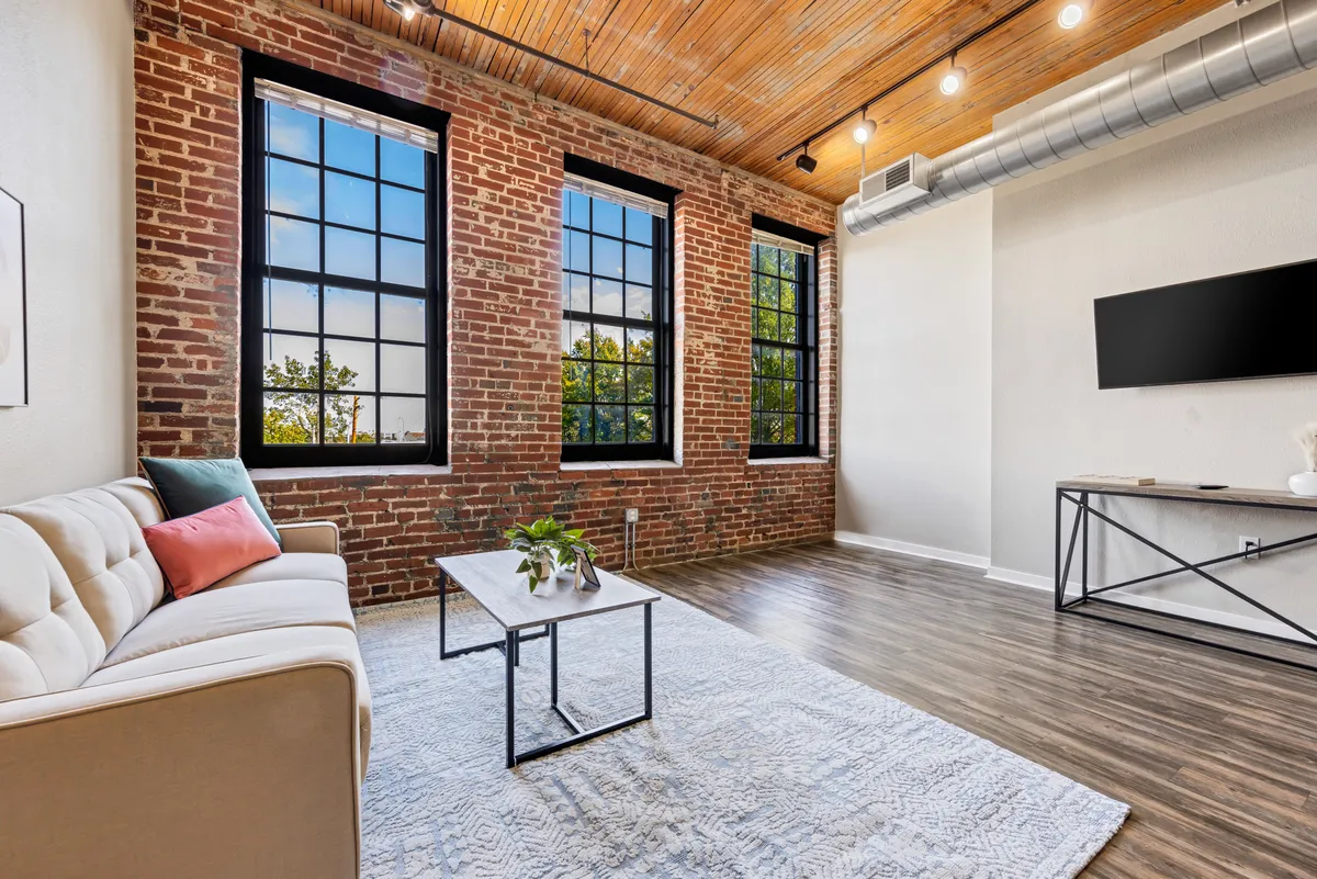 A bright, sunlit living room with comfortable seating, large windows framed by exposed brick walls, and a coffee table set for relaxation or socializing. The view through the windows hints at the city beyond, and the overall design merges contemporary comfort with historic character.