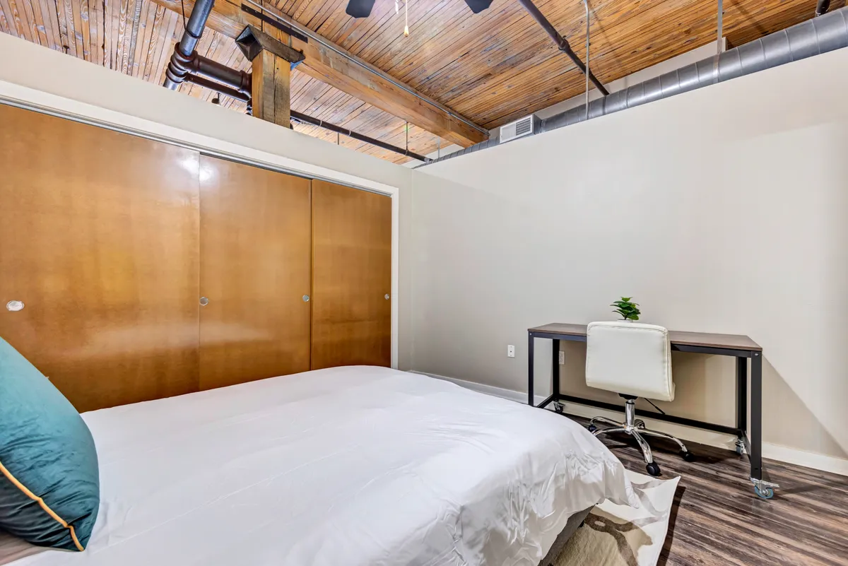 This image shows a serene bedroom with a plush bed placed beneath a wooden ceiling, accompanied by a dedicated desk space for working or reading. The atmosphere is peaceful, ideal for both relaxation and productivity. Natural light streams in, highlighting the tranquil setting and modern comforts.